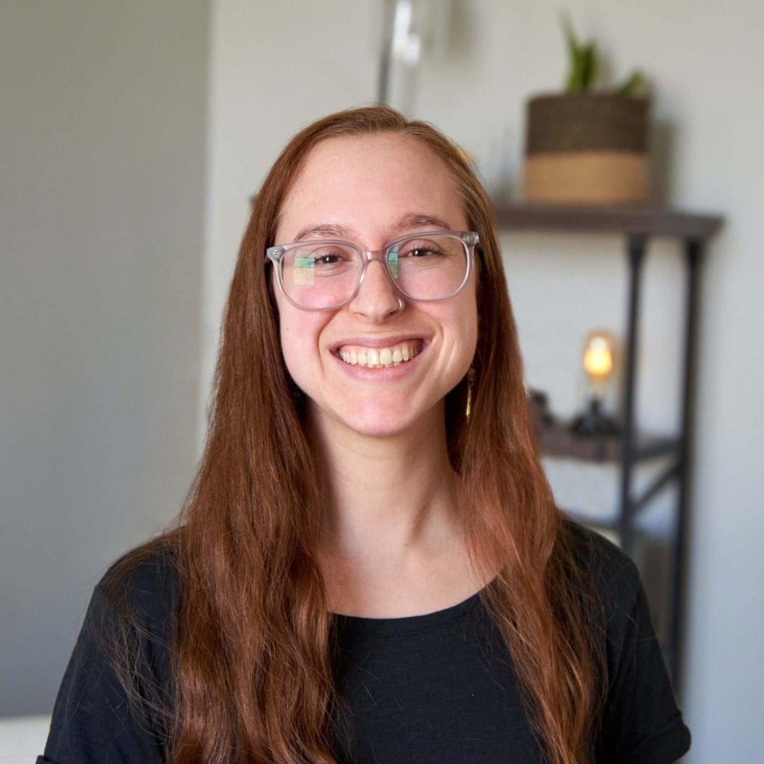 Caitlin Siekerka, therapist in Philadelphia with red hair and brown eyes. Caitlin is looking at the camera smiling in an office at The Better You Institute with a shelf and light in the background while wearing clear glasses and a black shirt.