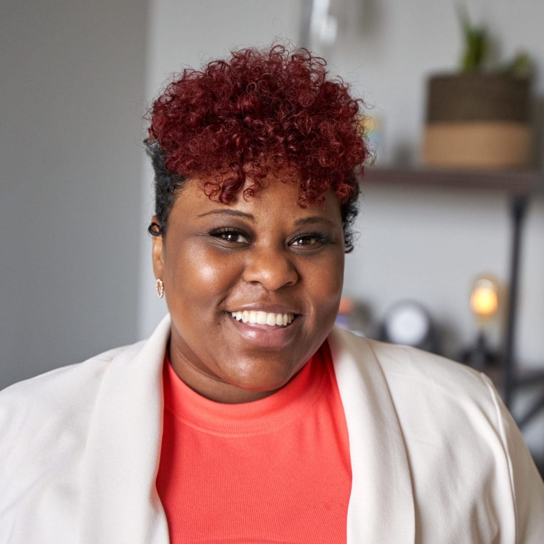 Willena “Lena” Hayes, therapist in Philadelphia with brown and red hair with brown eyes. Lena is wearing a coral shirt with a white blazers and is looking at the camera smiling in an office at The Better You Institute with a shelf and light in the background.