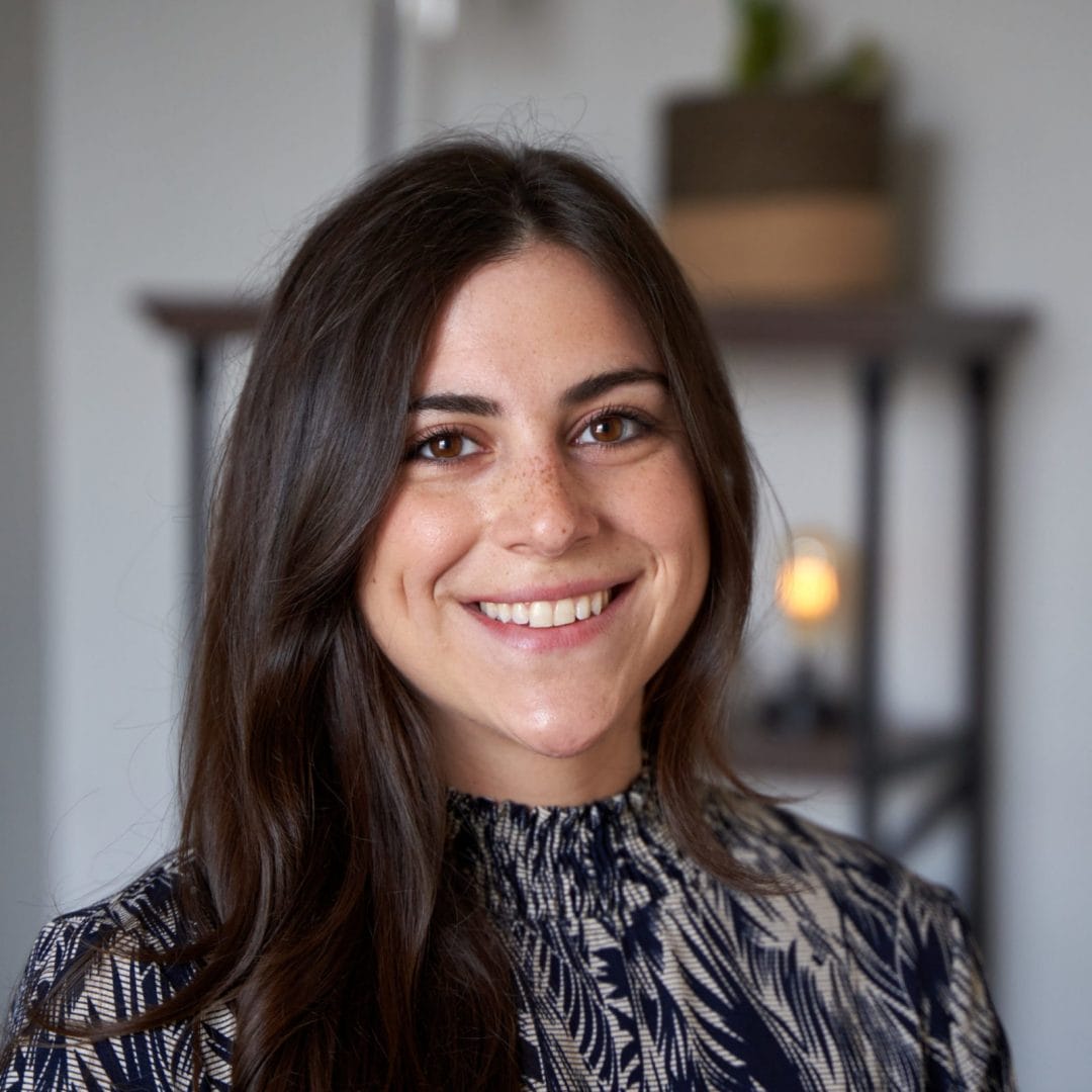Brianna Intili, therapist in Philadelphia with brown hair and brown eyes. Brianna is wearing a dark blue patterned shirt and is looking at the camera smiling in an office at The Better You Institute with a shelf and light in the background.