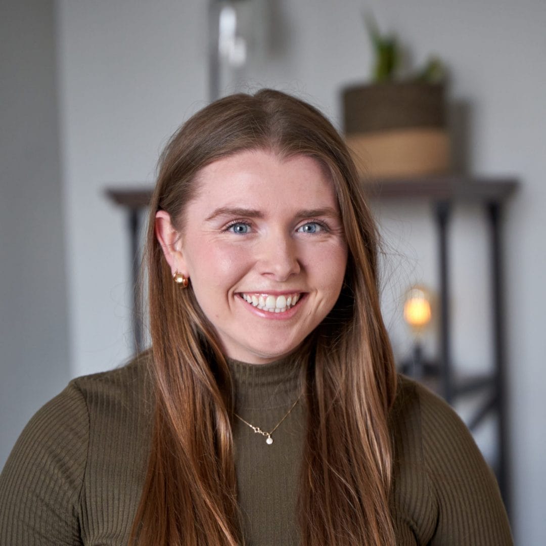 Claire Savage, therapist in Philadelphia with brown hair and blue eyes. Claire Savage is wearing dark green turtleneck shirt with gold earrings and necklace. She is looking at the camera smiling in an office at The Better You Institute with a shelf and light in the background.