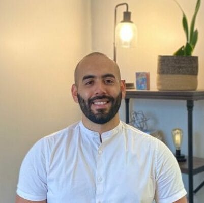 A man with a shaved head and beard, wearing a short-sleeved white shirt, stands smiling in a well-lit therapy office. Behind him, there's a bookshelf with plants and a lamp, creating a welcoming atmosphere that reflects Grant Morales's approach to systemic therapy with a focus on intersectionality, attachment, and trauma-informed practices.